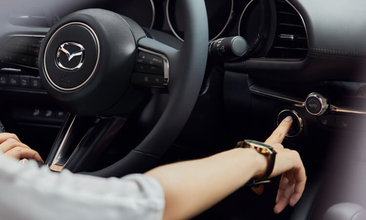 Close-up of steering wheel and front driver interior of Mazda3 Sedan, the driver’s hand is pressing the engine Start button. 