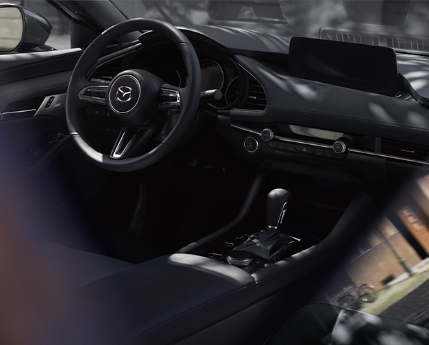 Close-up of steering wheel and front driver interior of Mazda3 Sedan, the driver’s hand is pressing the engine Start button. 
