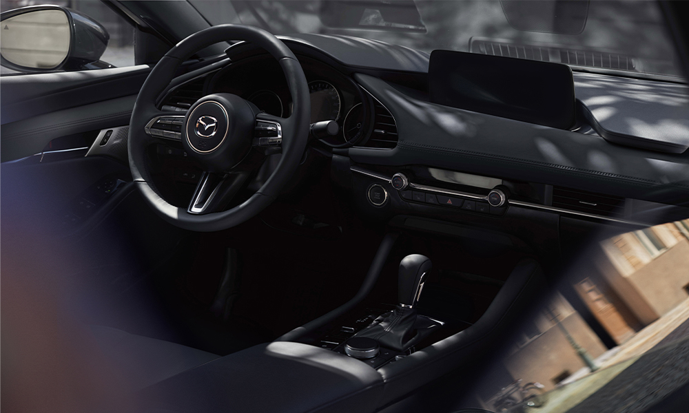 Close-up of steering wheel and front driver interior of Mazda3 Sedan, the driver’s hand is pressing the engine Start button. 