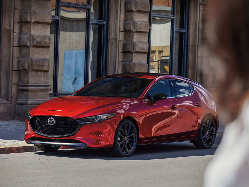 Soul Red Crystal Metallic Mazda3 Sport parked in front of city building. Grill is visible, sun and shadows play on contours of exterior.