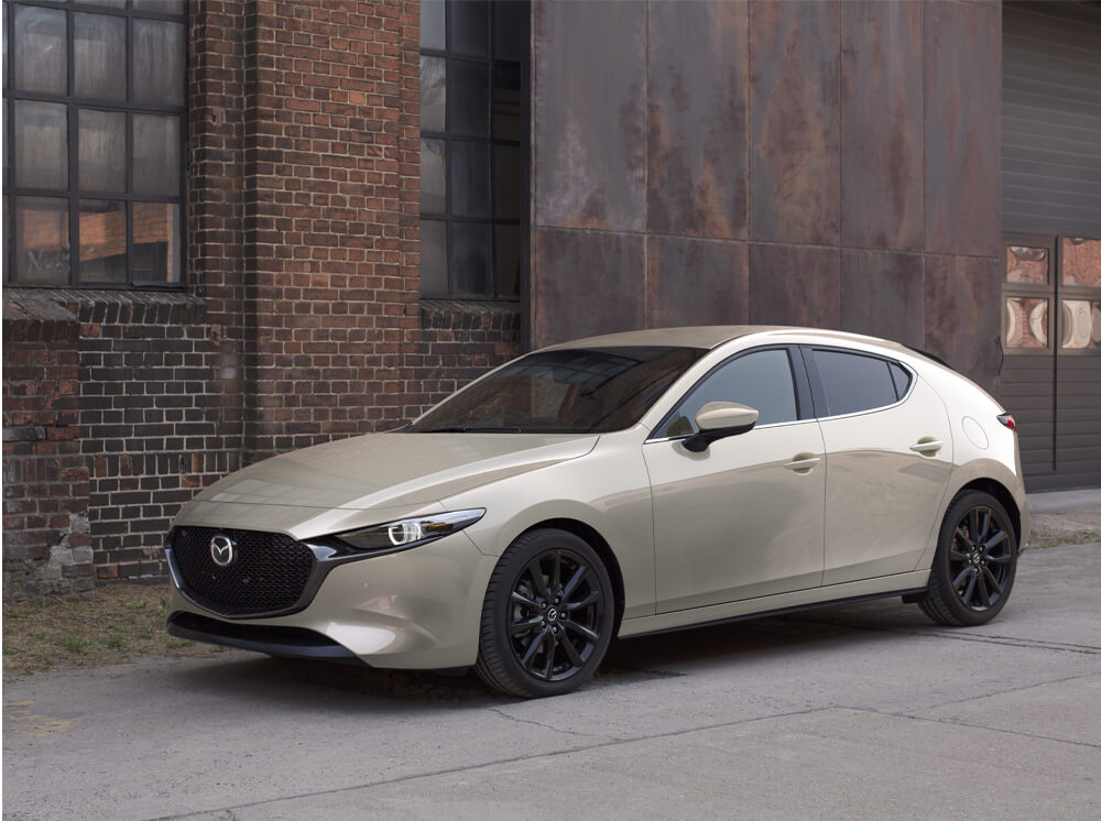 Suna Edition Mazda3 Sport parked in front of raw brick and steel plate industrial building in background. 