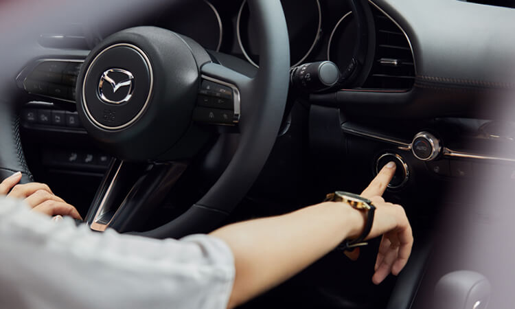 Close-up steering wheel and front driver interior of Mazda3 Sedan, the driver’s hand is pressing the engine Start button. 