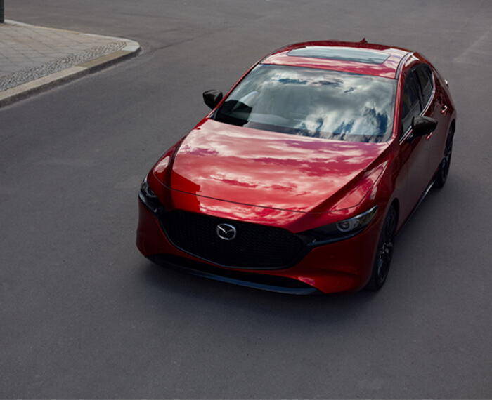 Above ¾ shot of Mazda3 Sport driving down city street, sky and clouds reflected in hood and windshield. 