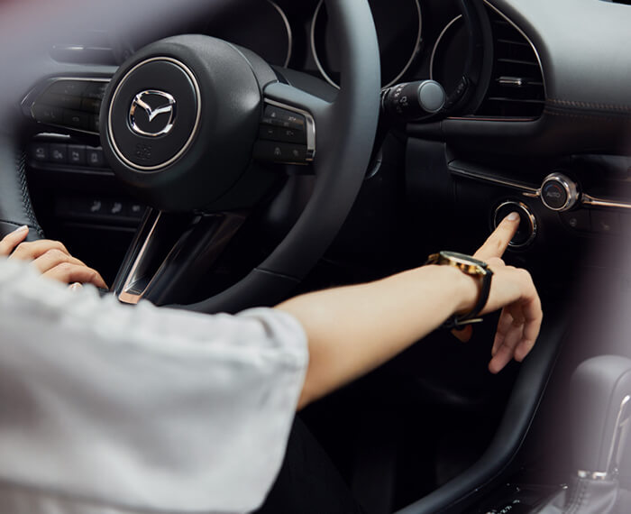 Close-up steering wheel and front driver interior of Mazda3 Sedan, the driver’s hand is pressing the engine Start button.