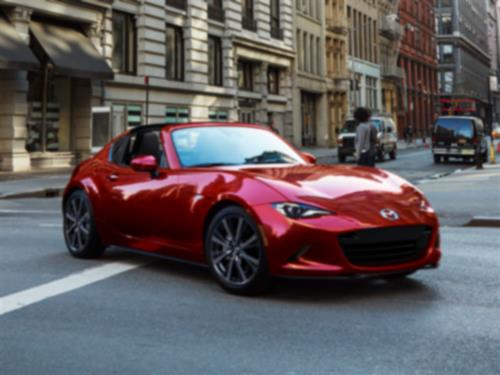 Vue aux 3/4 d’une MX-5 RF rouge qui descend dans une rue urbaine avec des bâtiments et des piétons en arrière-plan.  