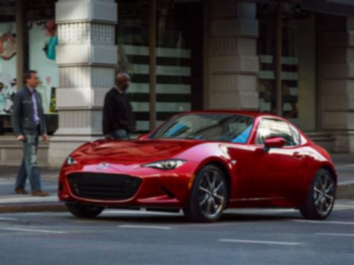 Une MX-5 RF rouge avec le toit fermé descend dans une rue urbaine avec des bâtiments et deux piétons visibles en arrière-plan.  