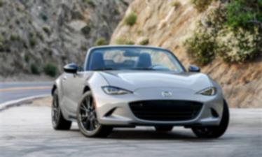 A silver MX-5 ST drives down a road cutting through the mountains.