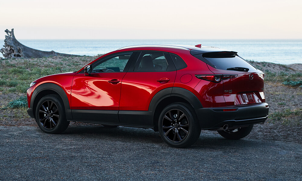 Vue latérale d'un Mazda CX-30 rouge métallisé Soul Red Crystal, stationné près d'un point d'eau sur un chemin de gravier.