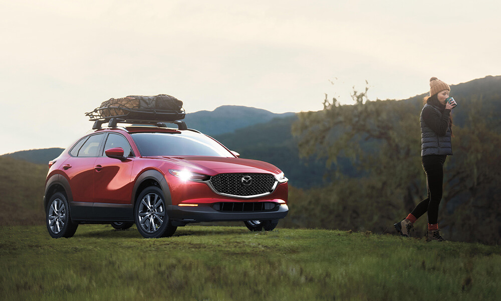 Une femme est debout derrière un Mazda CX-30 rouge vibrant cristal métallisé stationné dans un endroit architectural moderne avec des grandes fenêtes en vitre.