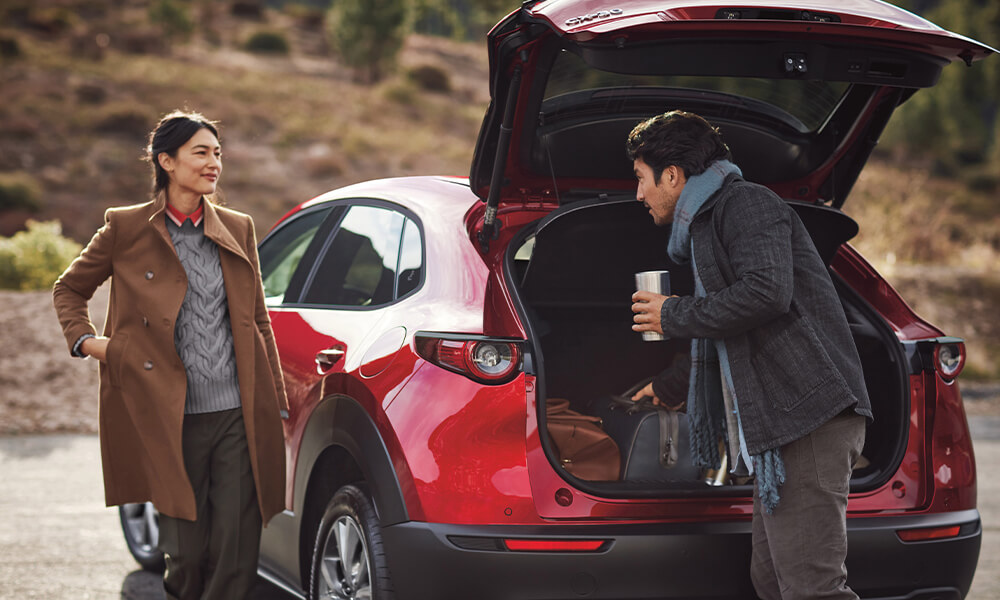 n homme et une femme se tiennent près du coffre d’un Mazda CX-30 rouge vibrant cristal métallisé avec un paysage extérieur flou en arrière-plan.