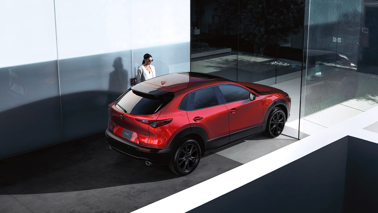 Une femme est debout derrière un Mazda CX-30 rouge vibrant cristal métallisé stationné dans un endroit architectural moderne avec des grandes fenêtes en vitre.