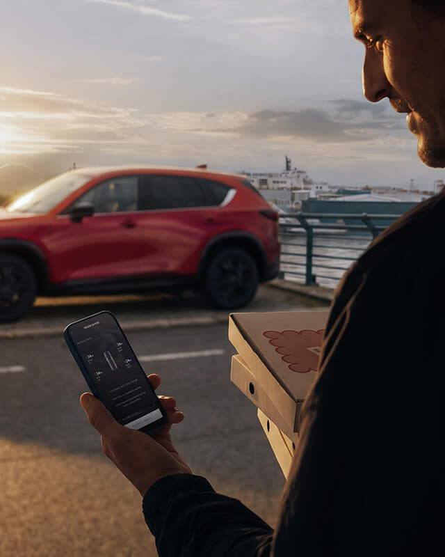 Man using his phone with a Mazda CX-5 parked near the waterfront in the background.