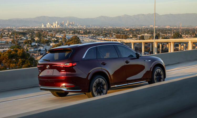 Side profile of a CX-90 driving on a highway with a city view in the background.