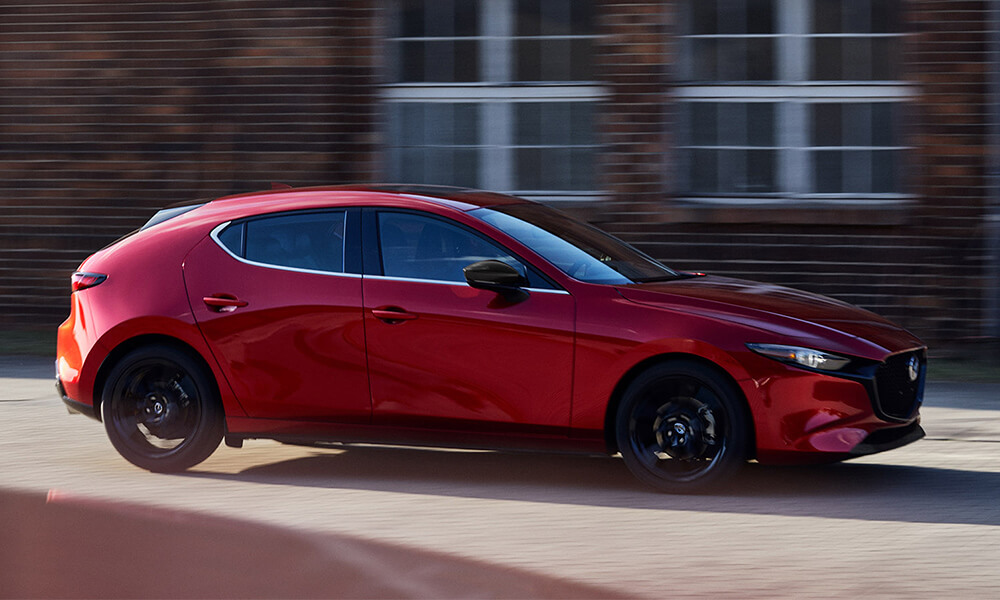 Vue de profil d’une Mazda3 Sport rouge vibrant cristal métallisé roulant à côté d'un immeuble flou.