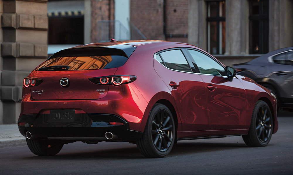 Une femme floue en avant-plan regardant vers une Mazda3 Sport rouge vibrant cristal métallisé montrée de profil.
