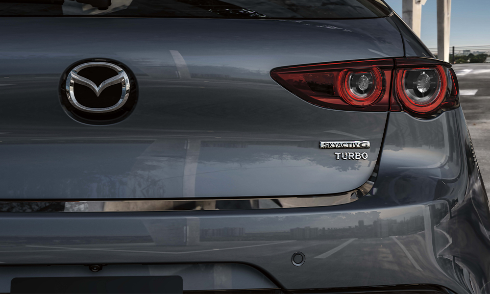 Vue de l’intérieur d’une Mazda3 Sport à travers le toit ouvrant panoramique, montrant une main posée sur le bouton de commande central.