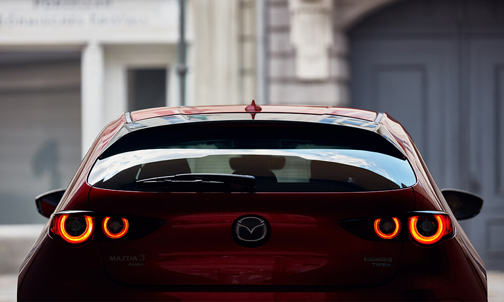 Vue de l’arrière d’une Mazda3 Sport rouge vibrant cristal métallisé avec des feux arrière illuminés.