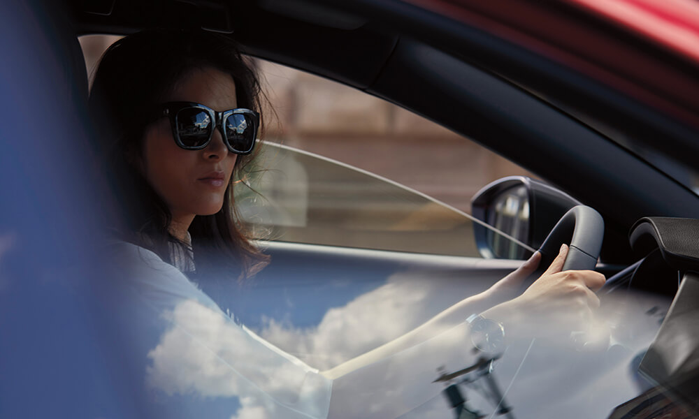 Vue de profil d’une femme portant des lunettes de soleil, à travers la fenêtre ouverte du côté passager, conduisant une Mazda3 Sport.