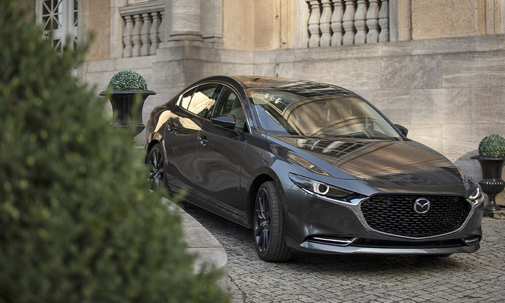 Front view of a Machine Grey Metallic Mazda3 with a potted ornamental bush in front and a mansion behind.